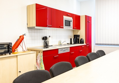 Im Vordergrund Tisch und Stühle, im Hintergrund eine rote Küchenzeile mit elektronischen Geräten